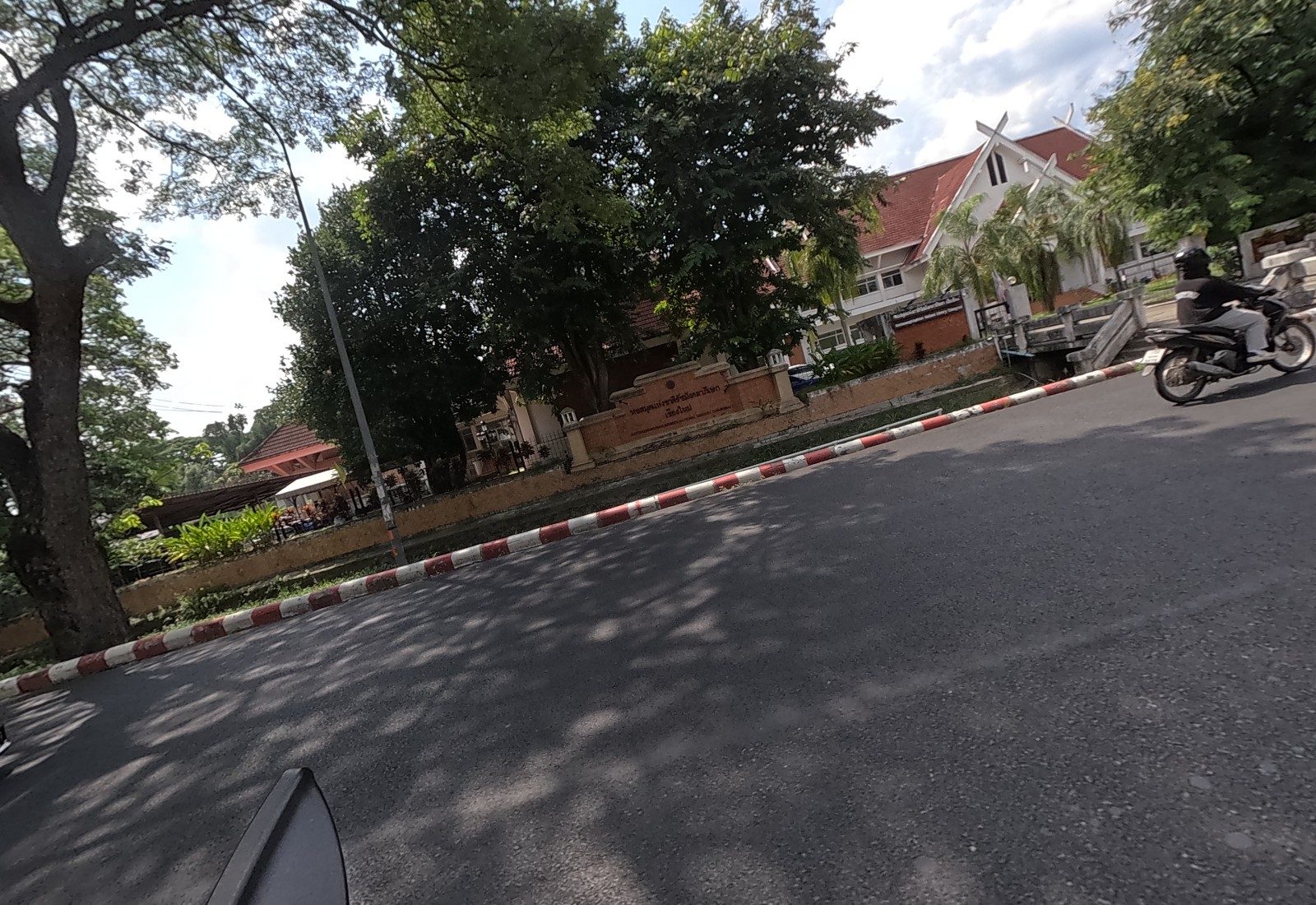 The brick name wall of the Ratchamangkhalaphisek National Library stands before its red-roofed Lanna buildings; a motorcyclist leaves the gate.