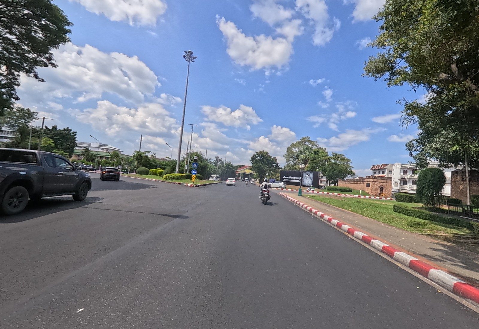 The road nears the Suan Dok Gate junction: the brick gate and wall at right, a black memorial board with a royal portrait, and a traffic island with keep-left signs.