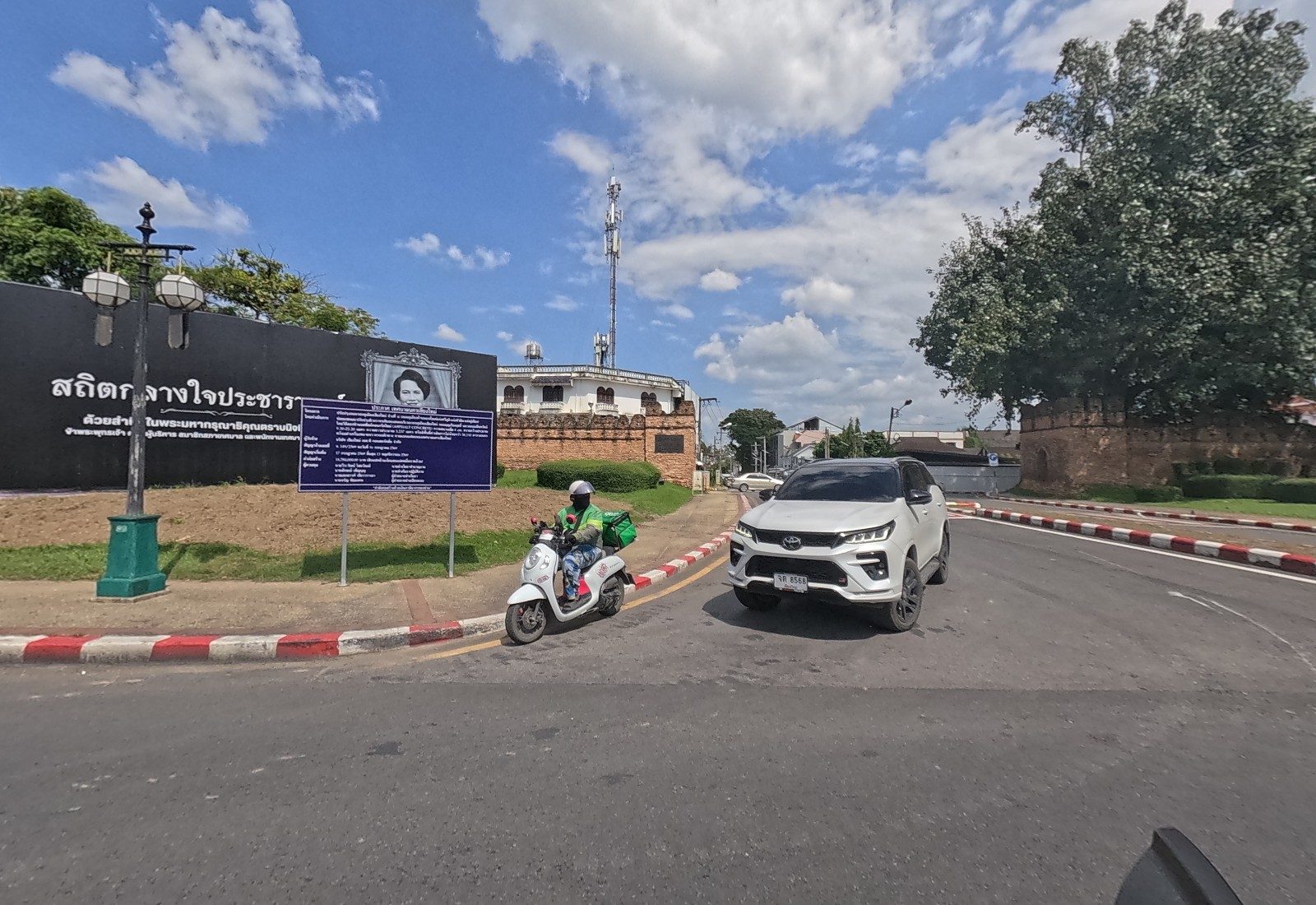 The memorial board with a royal portrait and a blue municipal notice stand before Suan Dok Gate; a food-delivery rider and a white SUV come out of the gate road.