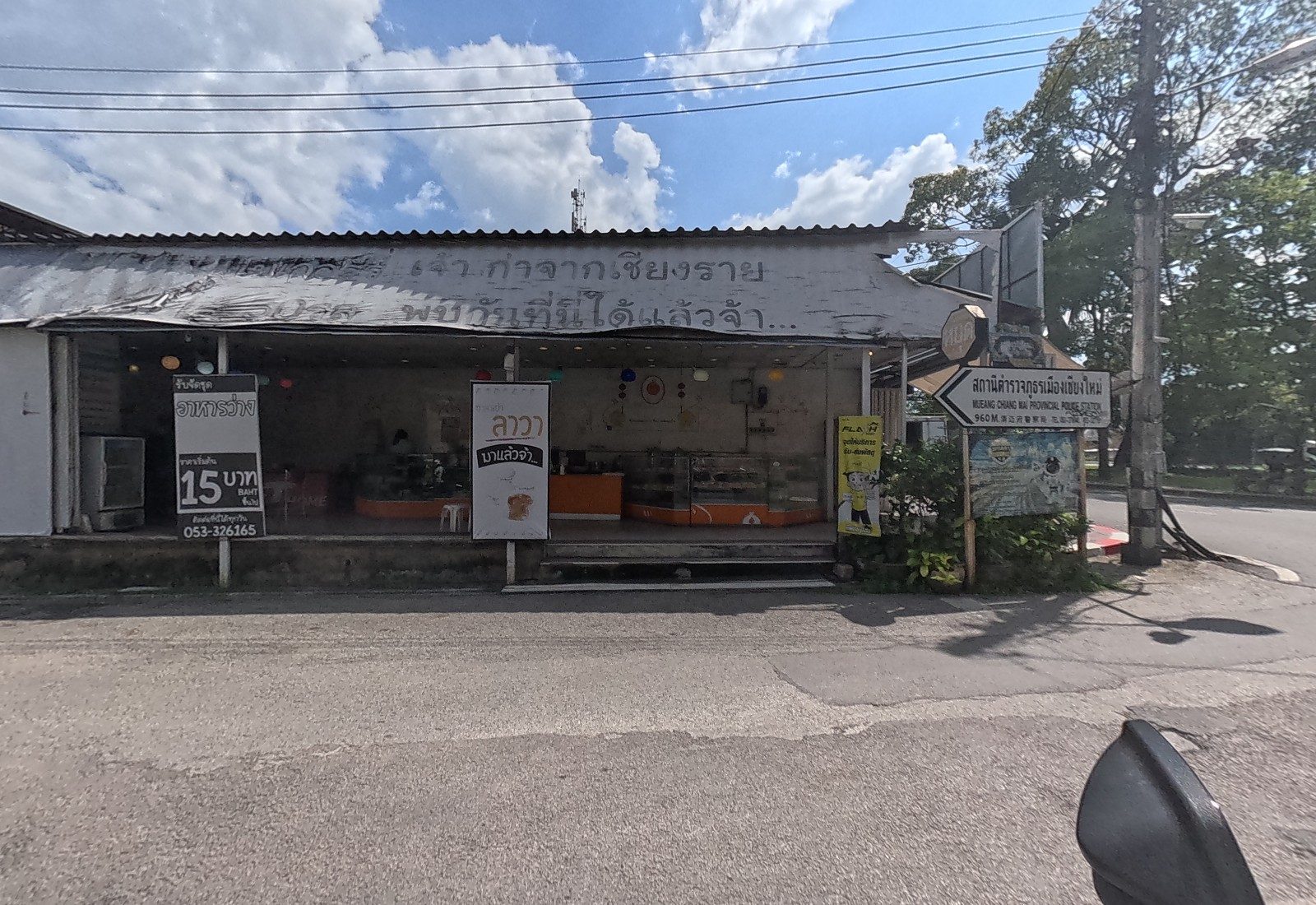 The snack shop's open front under a hand-painted roof, with 15-baht and lava-bun boards, a Flash parcel sign and the police-station direction sign.