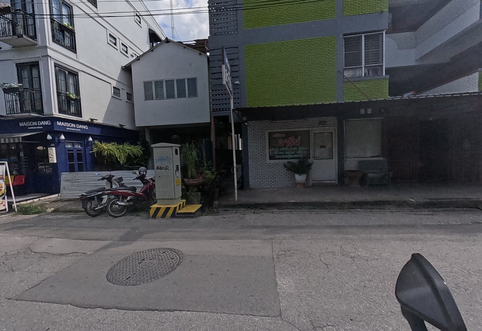 Maison Dang boutique hotel's navy frontage with a menu board sits beside a small white house and the อารีย์ salon behind a glass door under the green building.