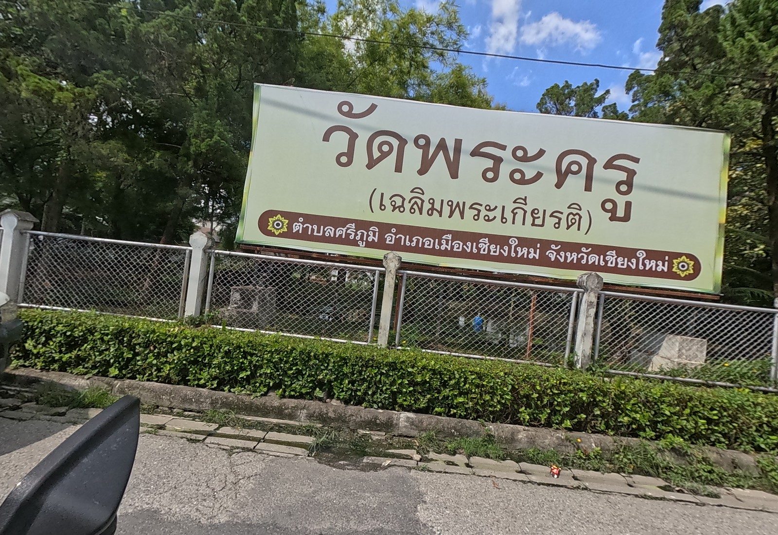 A large sign for วัดพระครู (เฉลิมพระเกียรติ), ตำบลศรีภูมิ อำเภอเมืองเชียงใหม่ จังหวัดเชียงใหม่, stands behind the chain-link fence and hedge.