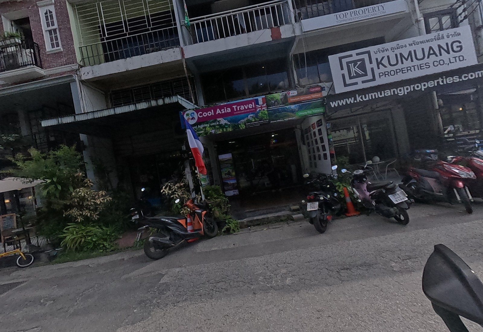 Cool Asia Travel's agency front, with Elephant Sanctuary Thailand and Chiang Mai Trekking panels and a French flag, sits under the Kumuang Properties and Top Property signs; motorbikes park in front.
