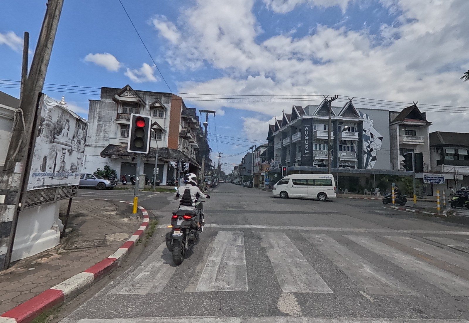 At a red light: the August Hostel with its geometric lion mural faces the junction, a white van crosses and a motorbike waits on the zebra crossing.