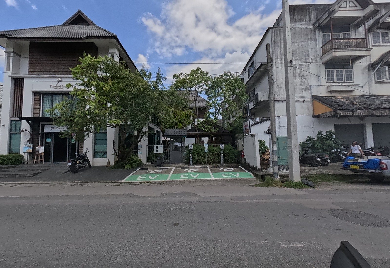 The Paraiso Hotel's white-and-timber front, with three green EV charging bays and chargers beside its gate; a woman walks past parked motorbikes by the old building at right.