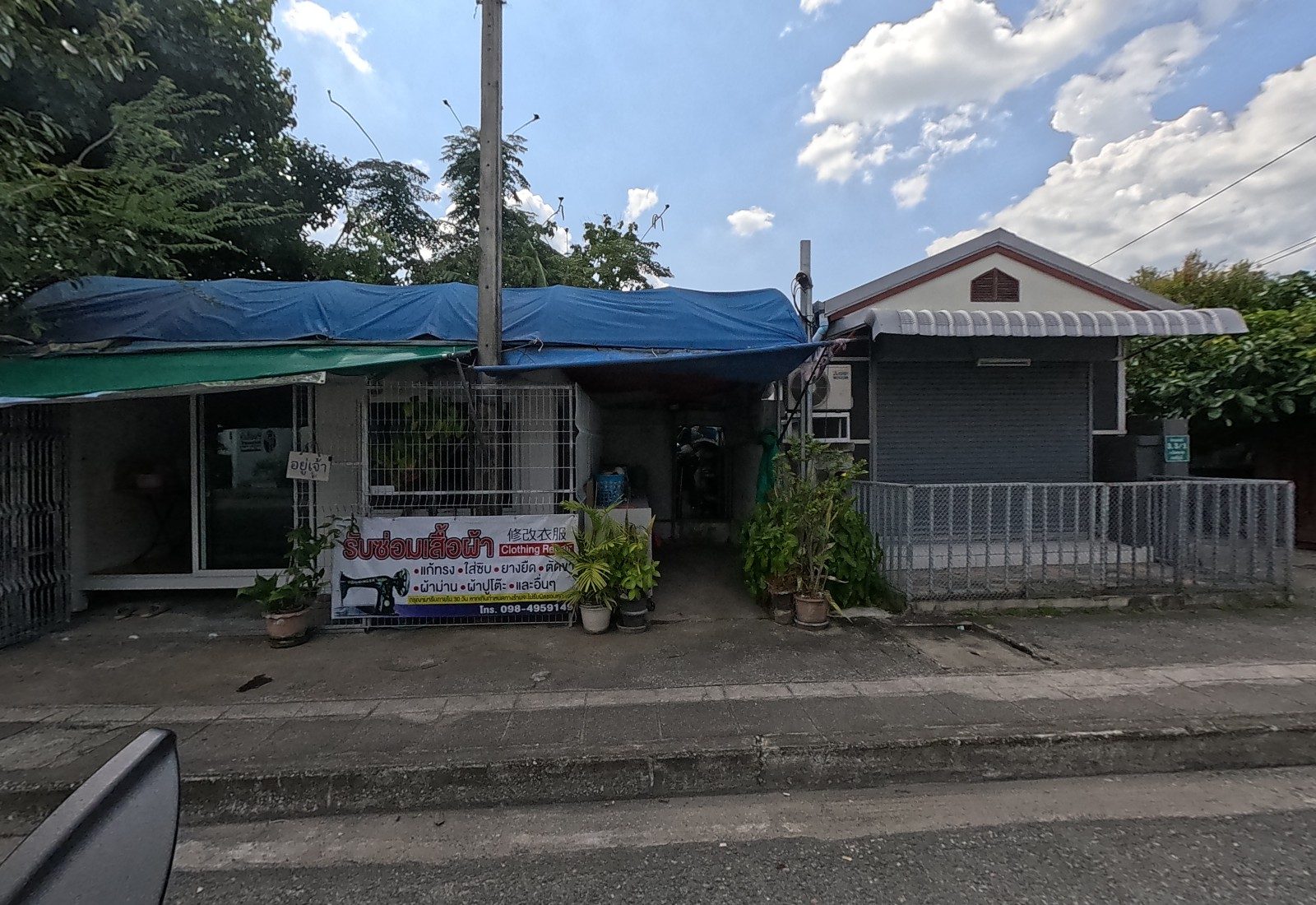 A clothing-repair shop under a blue tarp roof with a banner รับซ่อมเสื้อผ้า, 修改衣服, Clothing Repair, and an อยู่เจ้า sign on the door; a small shuttered grey house stands next to it.