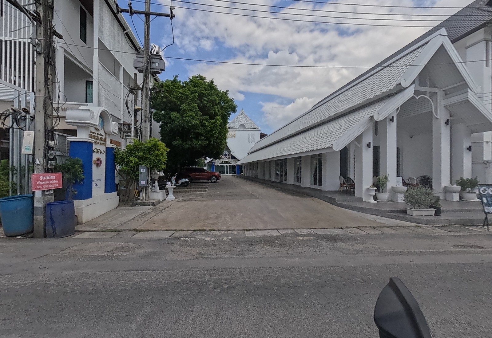 A driveway leads between the blue-and-white gate pillar of the SINGHARA… boutique hotel and a long white Lanna-roofed arcade to a car park and a white hotel building; a red sign on the pole advertises a housekeeper vacancy.
