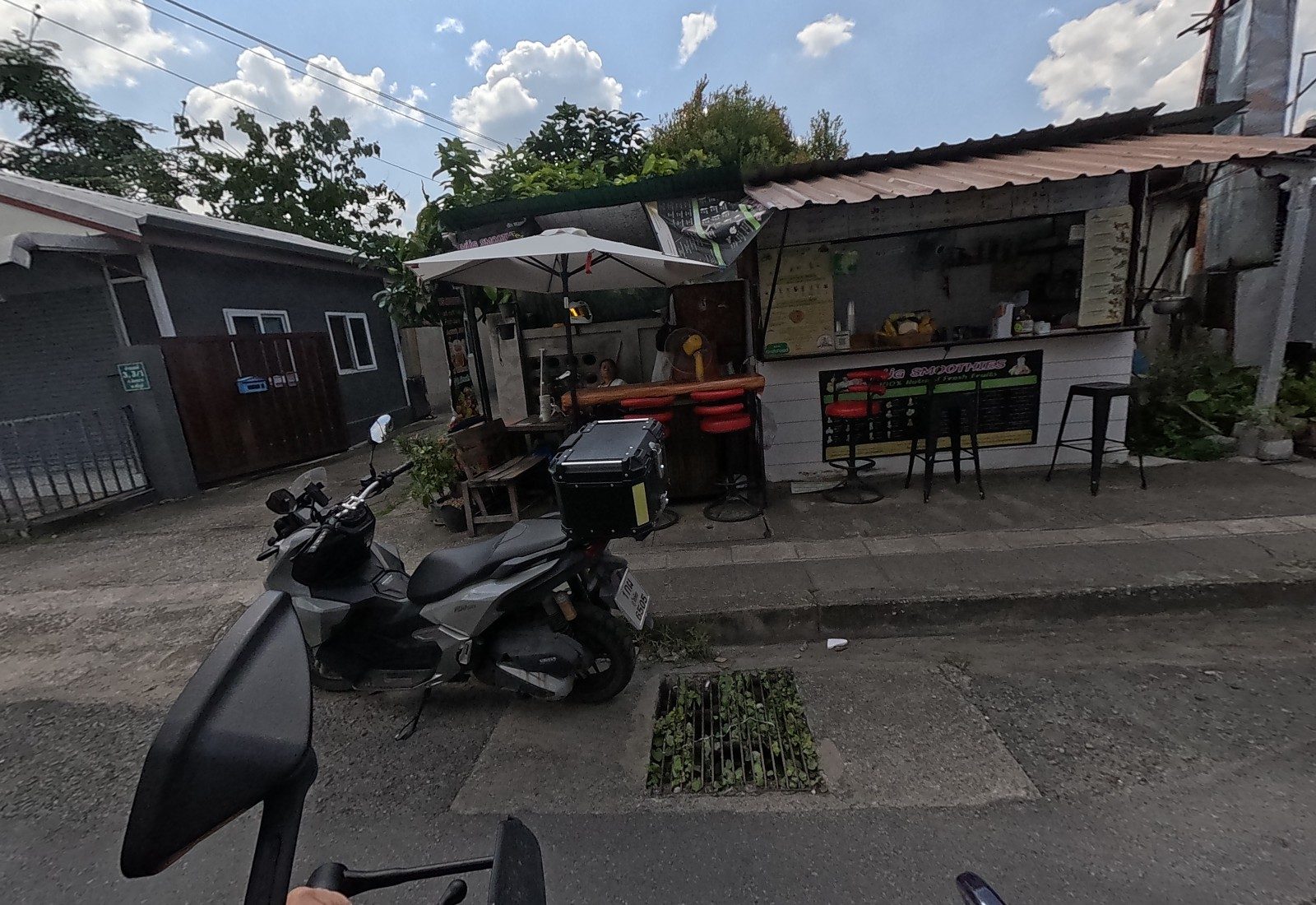 A smoothie kiosk with red bar stools, an umbrella and a chalk menu board, with a woman inside; a touring scooter parks in front.