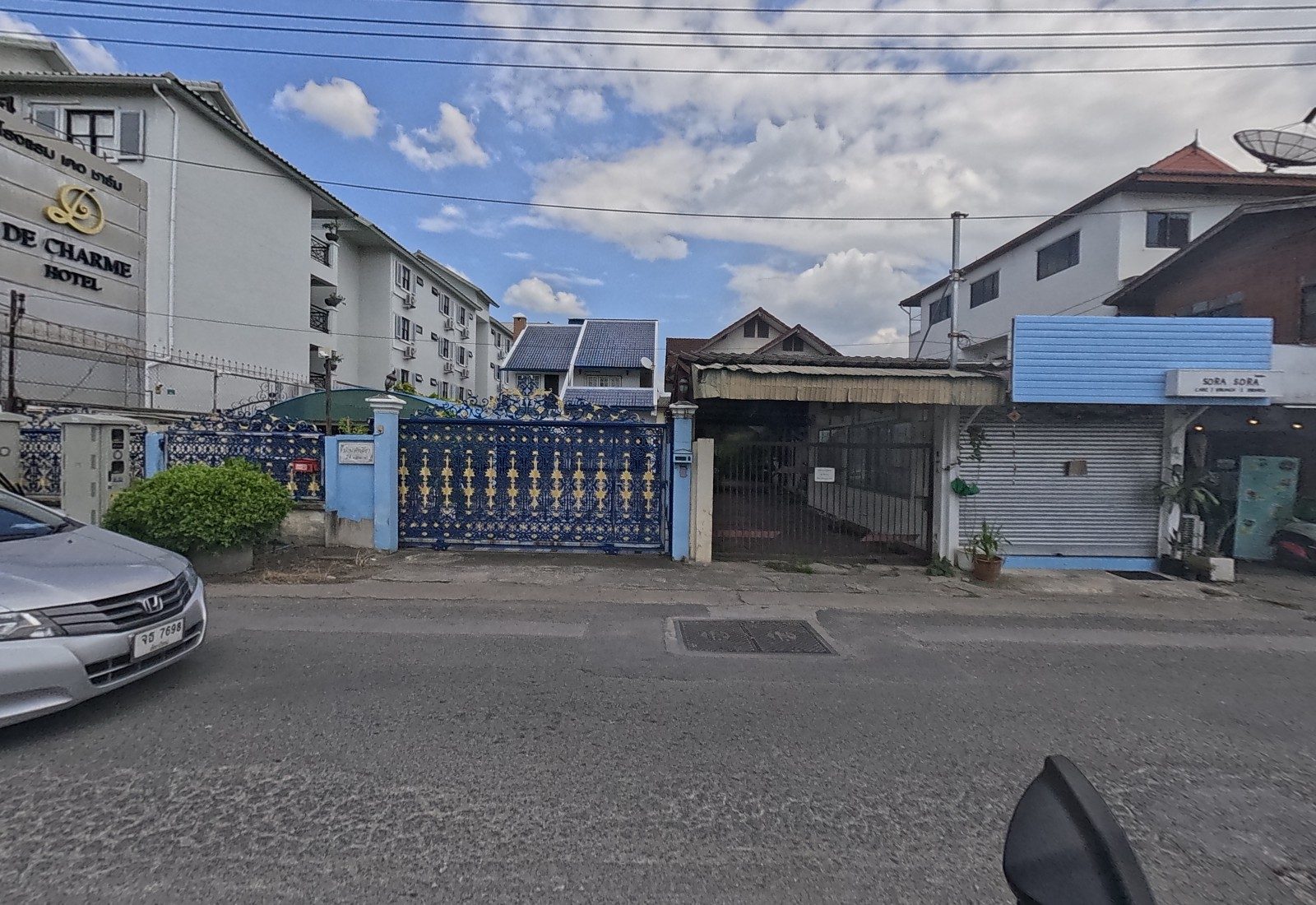 The De Charme Hotel's sign and four-storey block stand beside an ornate blue-and-gold gate, an old house behind a steel grille and the shuttered SORA SORA café.