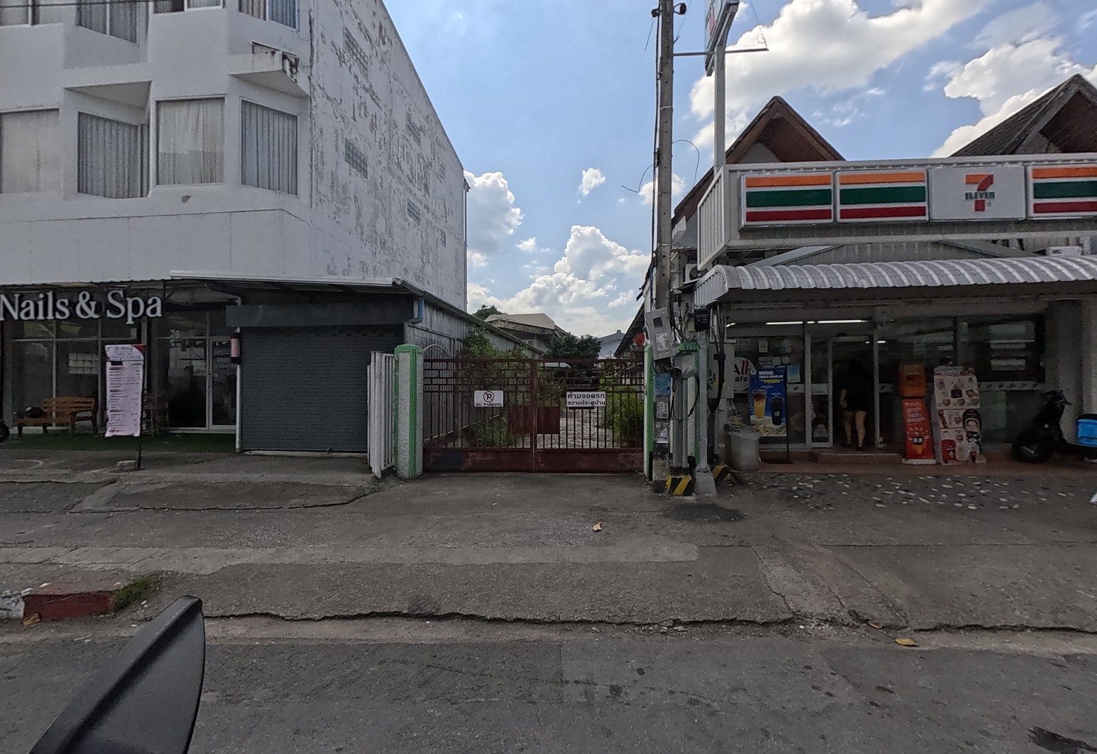 A locked gate with no-parking signs sits between K-perfect Nails & Spa and a 7-Eleven with an All Cafe counter; a woman walks into the shop.