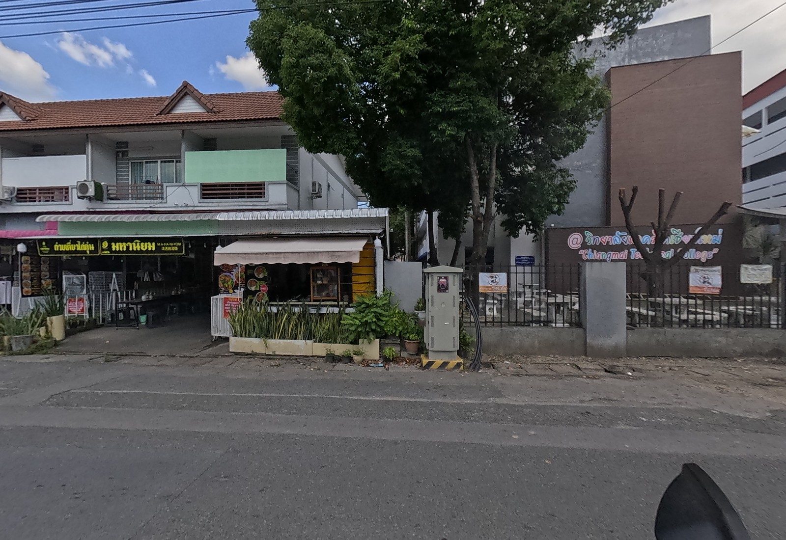 Across the road, the Maha Niyom stewed-chicken noodle shop and a small kitchen kiosk with a yellow wall sit beside the college fence and its neon lettering.