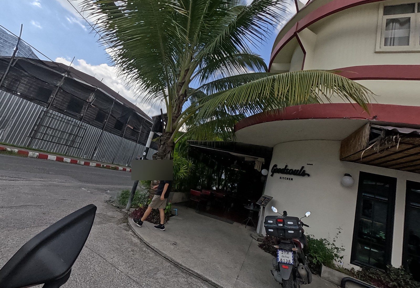 Goodsouls Kitchen's curved cream-and-red corner with a palm; a man walks past a delivery scooter.