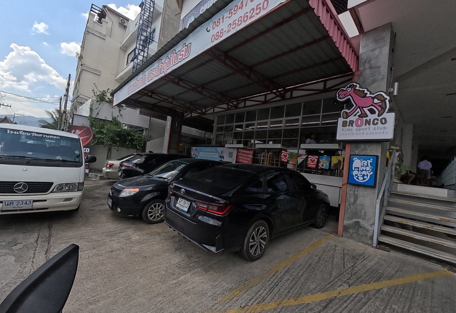 Ground floor of the gym building: the Bronco Kids sport club sign, an Art and Play light box and ice-cream flags above parked cars.