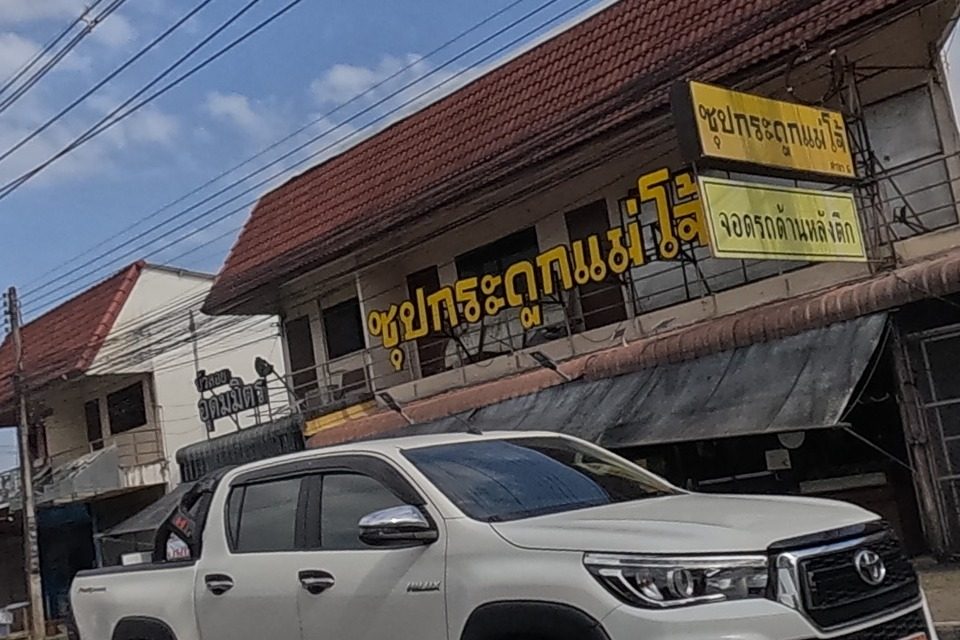 A row of shophouses with a large red sign reading Sup Kraduk Mae Jo (bone soup restaurant) and a yellow parking notice above it, a white pickup passing in front.