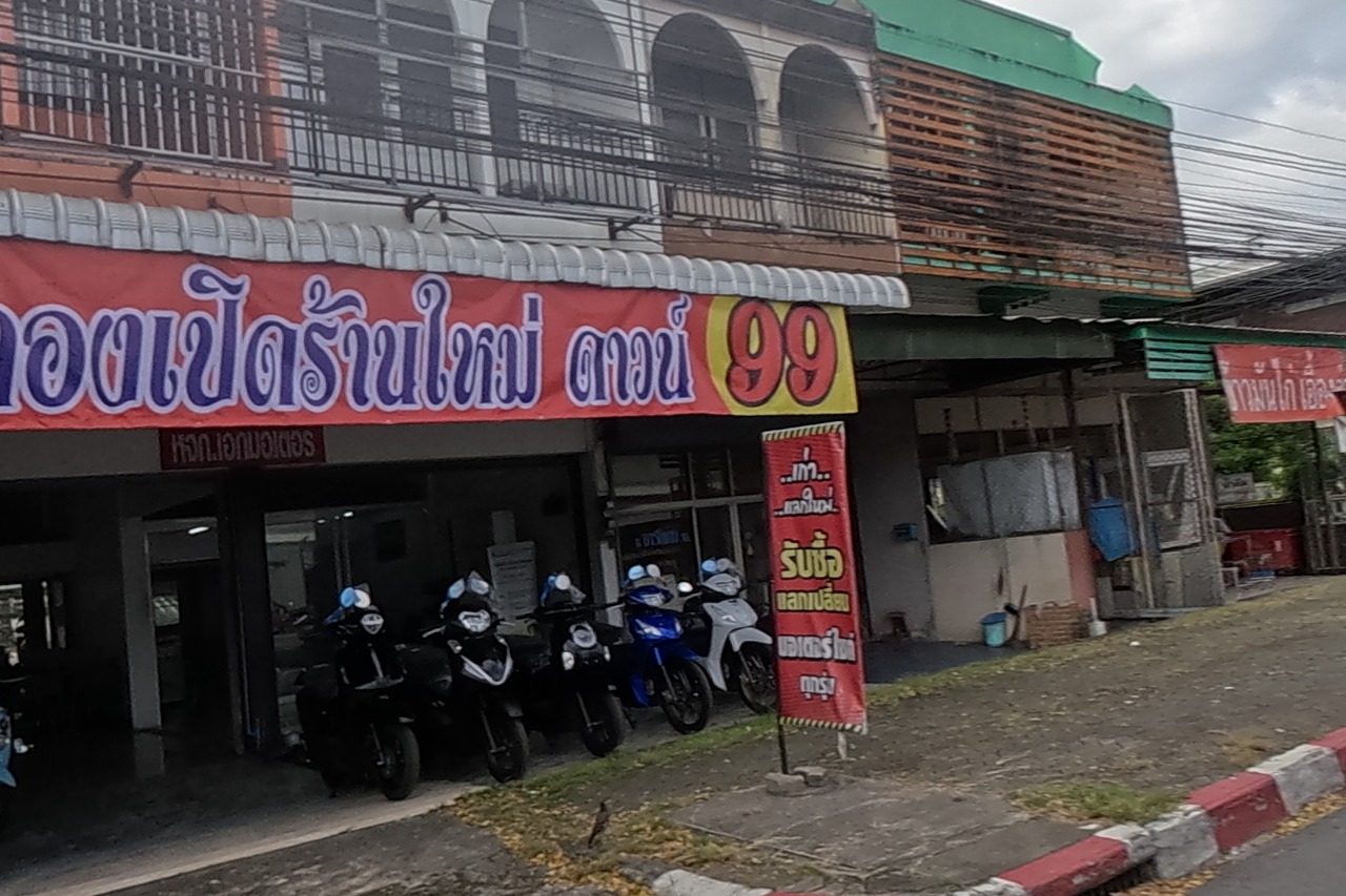 Red banners advertise a new motorbike shop with a 99-baht down payment promotion and buy/trade-in service, motorbikes lined up under the arcade; the road continues past more shophouses.
