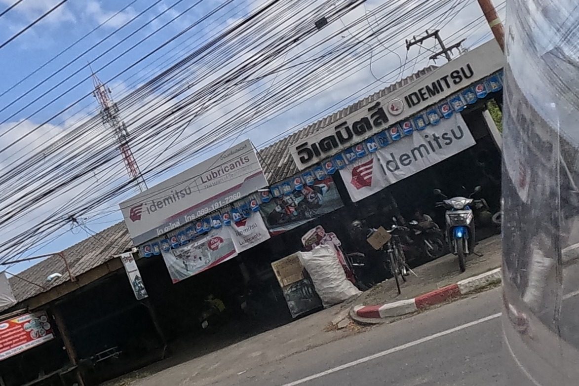 Across the road, a motorcycle repair and lubricant shop with large Idemitsu branded signage and a phone/LINE number, motorbikes parked inside.