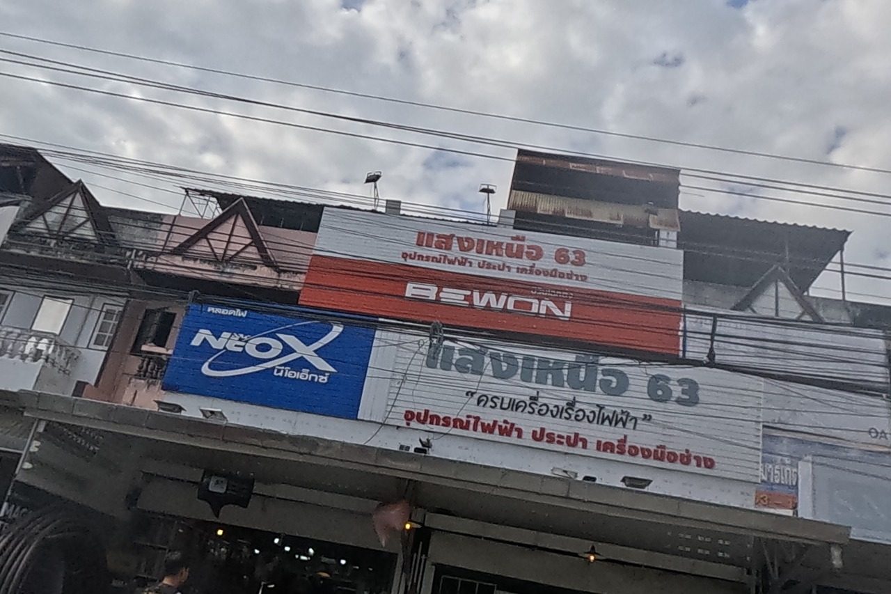A large hardware store called Saeng Nuea 63 selling electrical, plumbing and tool supplies, with NEO-X and BEWON brand signs, and a small tyre-repair stall beside it with a red banner.