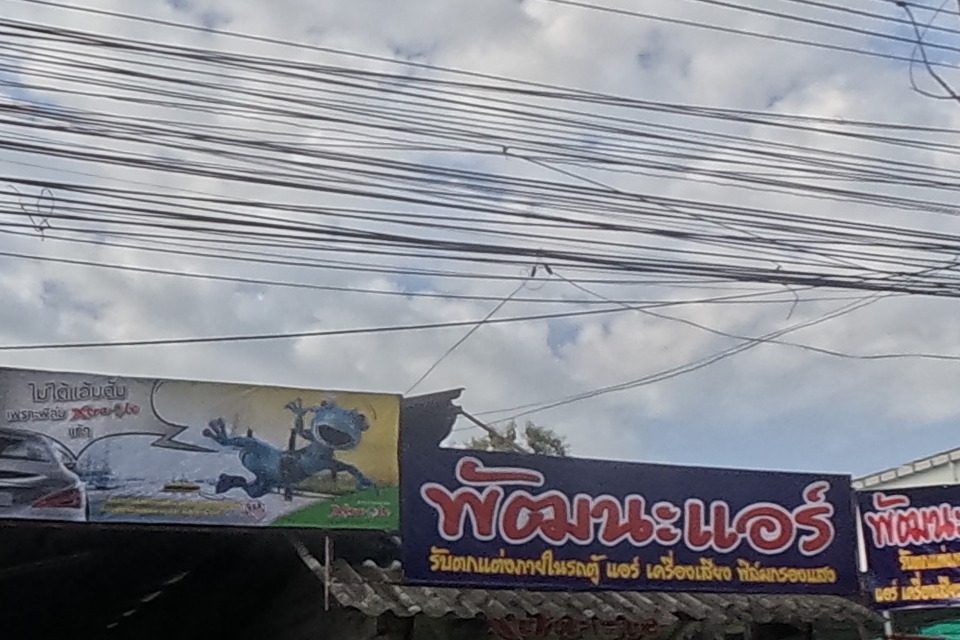 A car audio, air-conditioning and window-tint shop called Pattana Air, next to a fenced building with a red banner advertising the land and building for sale with a phone number.