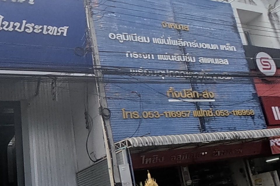 A large warehouse building carries a blue sign for a wholesale/retail liquor company, next to another shop selling aluminium, polycarbonate sheet and glass with a phone number; a small gilded spirit house sits at the aluminium shop's entrance.