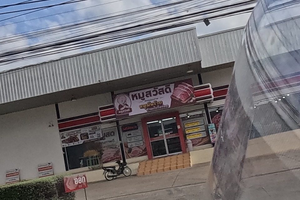A fresh pork shop with a red sign and pig logo sits beside a 7-Eleven and a blood-test clinic sign, in a strip of shops set back from the road.