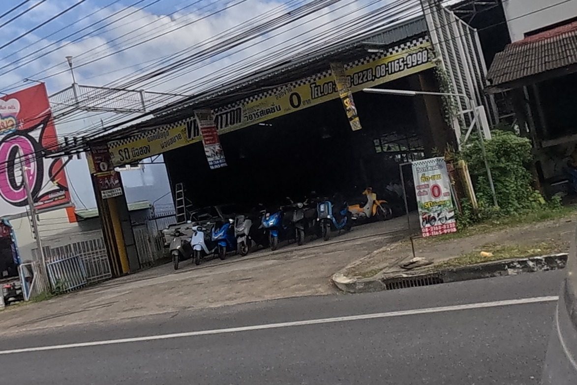 A used-motorbike rental/sale yard with scooters lined up under a shed, a large yellow banner reading VR Station and 0-baht promotional pricing, with phone numbers.