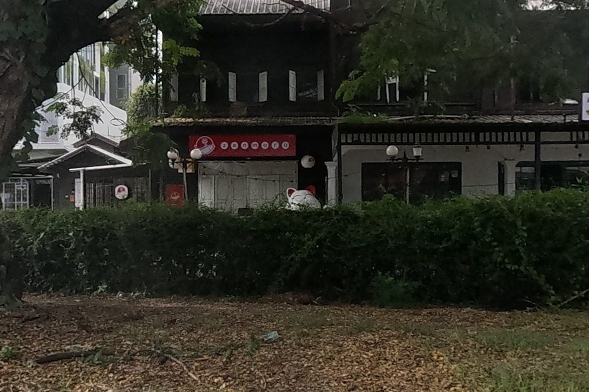 An old dark wooden two-storey Chinese-style shophouse with a red Zenmaru restaurant sign sits behind a hedge with a giant white lucky-cat statue peeking over the top, under a huge shade tree.