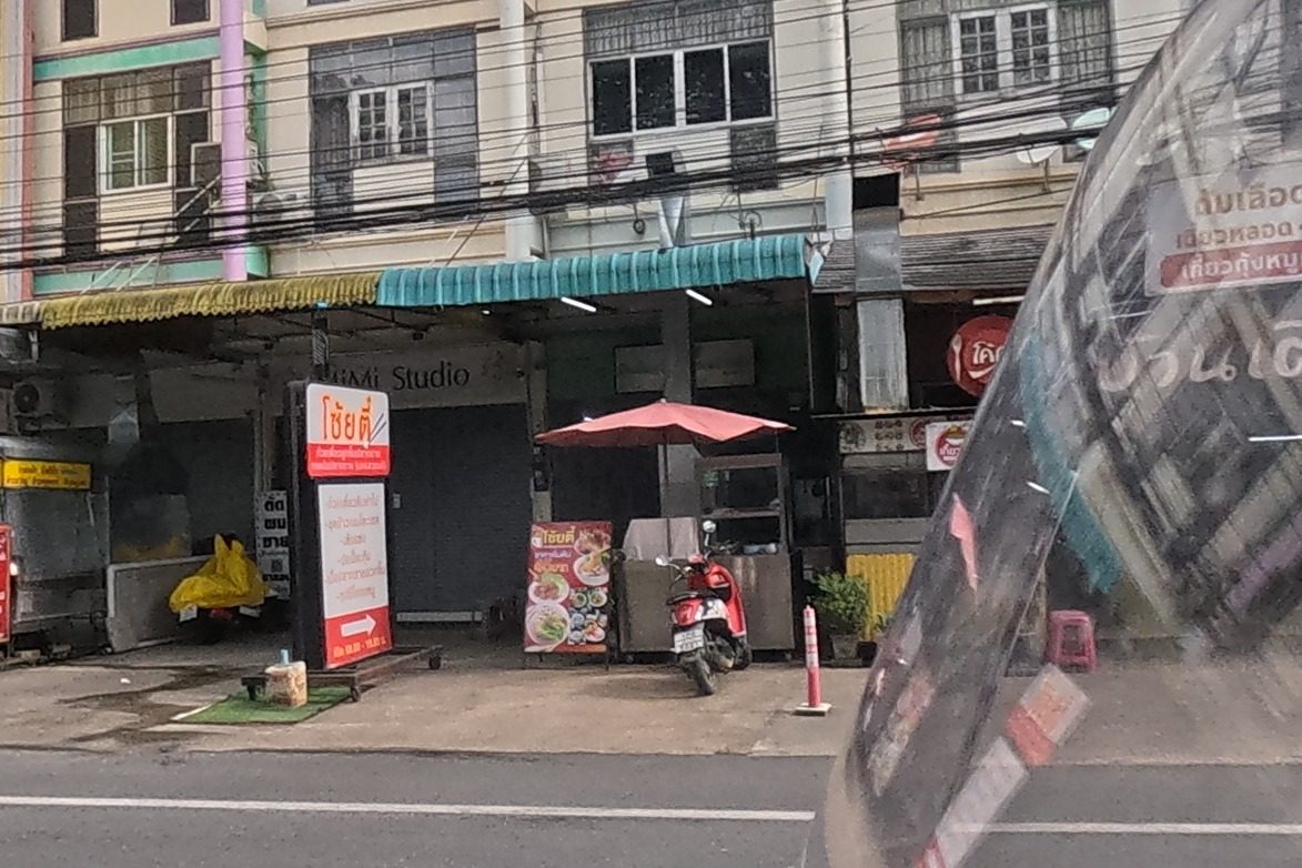 A row of colourful three-storey shophouses includes a restaurant called Baan Toem Im with a red menu sign listing blood soup and noodle dishes, next to a photo studio and other shops.