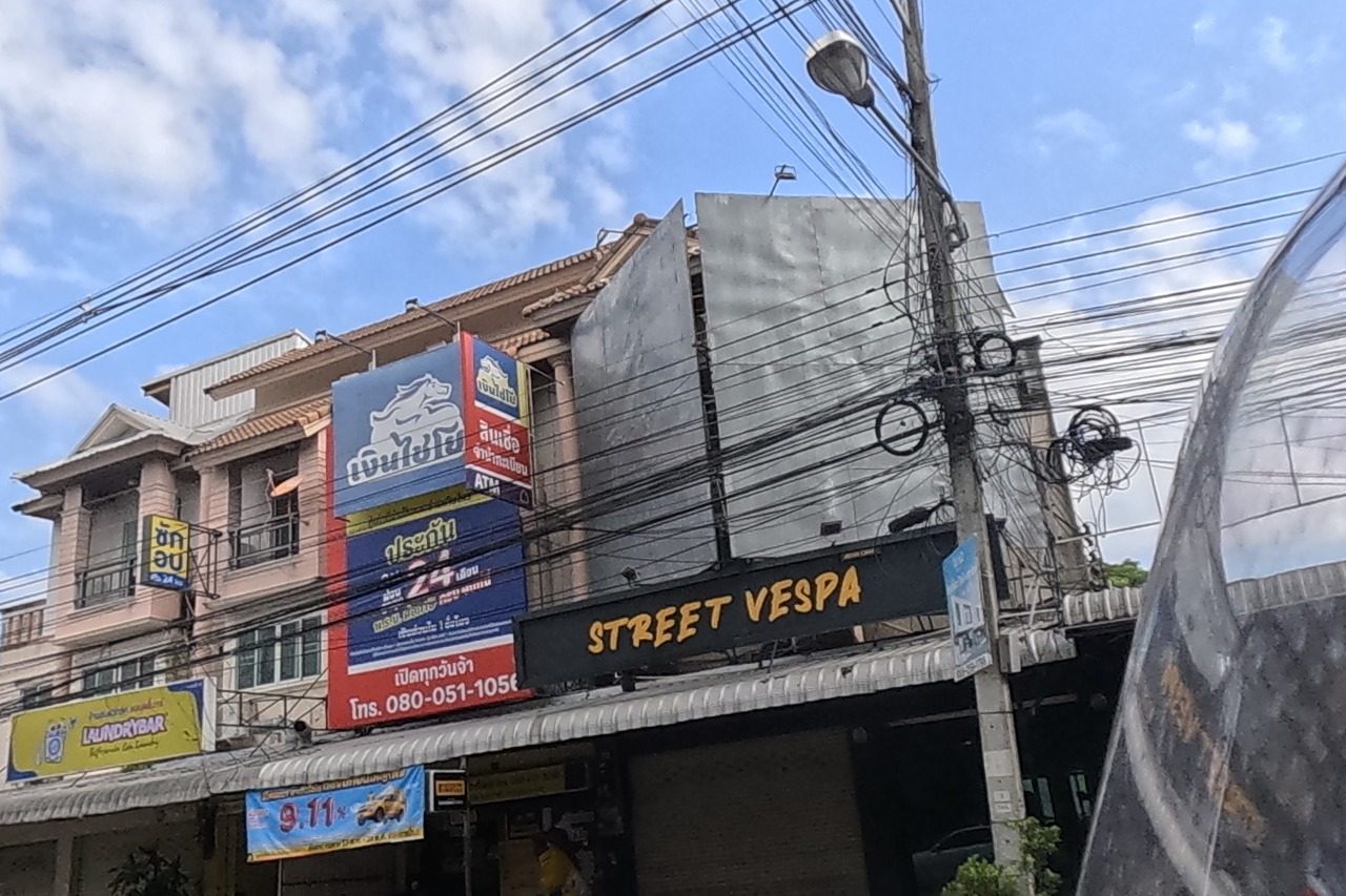 A three-storey building holds a Vespa scooter shop called Street Vespa, a pawn/loan shop called Ngoen Chaiyo advertising 24-hour service and ATM, and a Laundrybar self-service laundry.