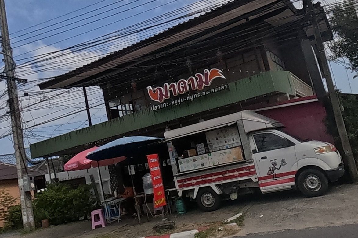 A seafood and hotpot restaurant called Madam with a red sign advertising grilled fish, mussels and moo-kratha sits beside a food truck; a green cannabis-shop sign is visible further along.