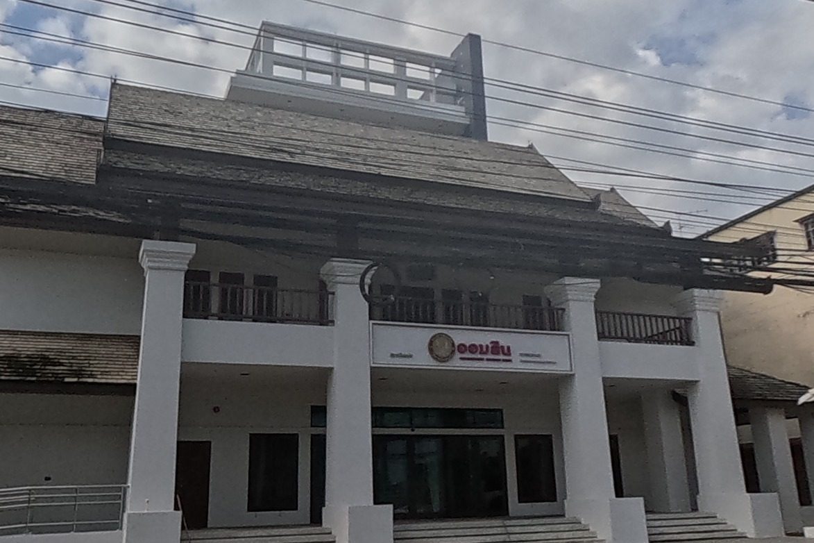 A grand Lanna-style white building with a red Government Savings Bank sign stands beside the brick-walled compound with its white chedi, a dark high-rise visible behind.