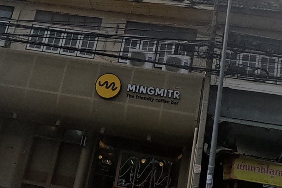 A row of old three-storey shophouses includes Mingmitr, a friendly coffee bar with a yellow circular logo, next to a red pawn-shop sign and other ground-floor shops with scooters parked outside.