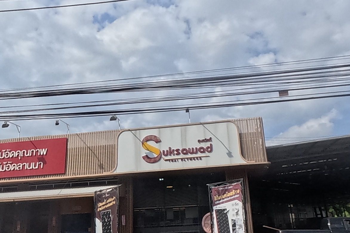 Full frontage of the 'Suksawad Thai Plywood' shop with its large red sign, pickup trucks parked outside.