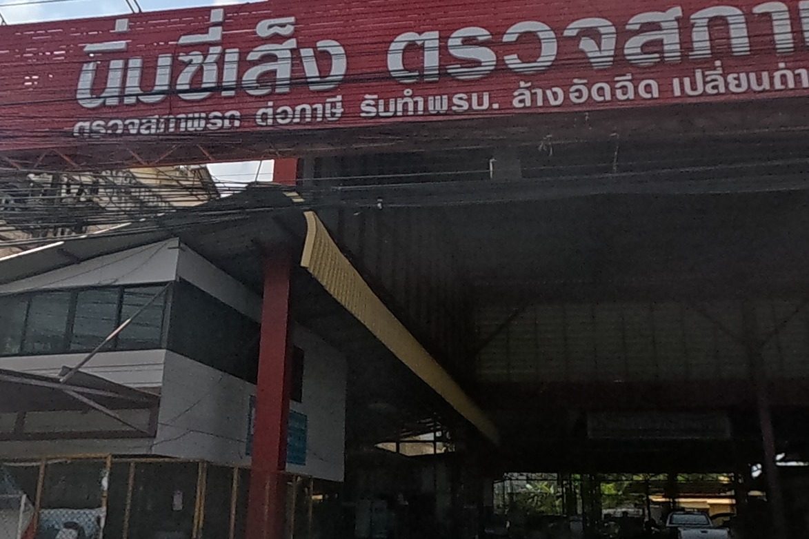 A large red-signed vehicle inspection centre, 'Nim Si Seng', offering inspection, tax renewal, insurance, car wash and oil change, with a hiring sign.
