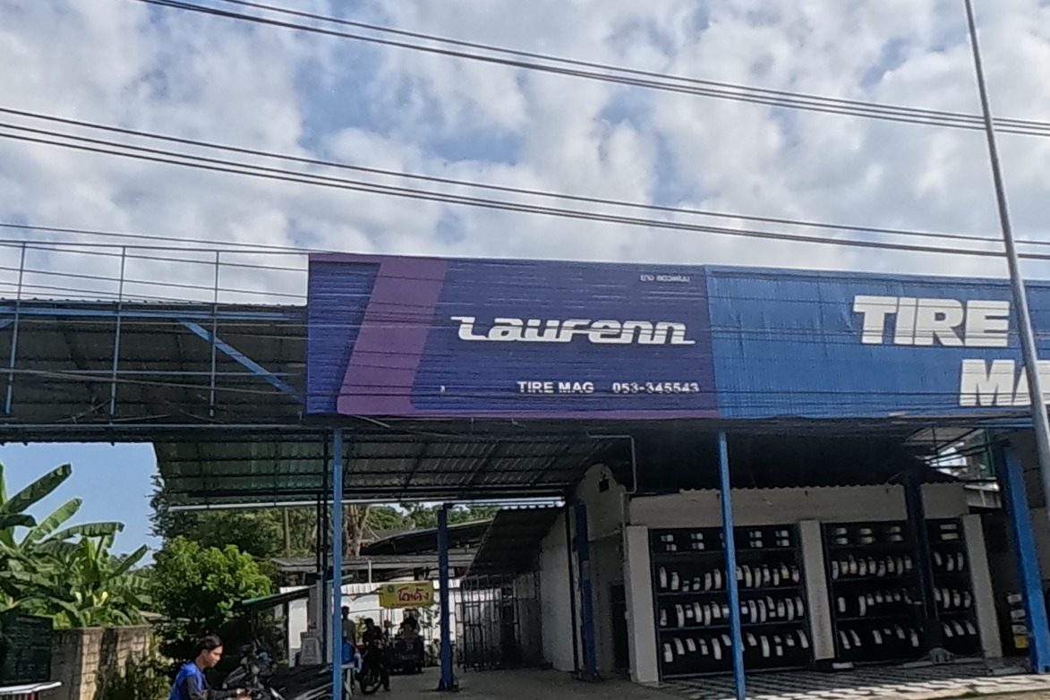 A roadside tyre shop with a blue Laufenn Tire Mag sign faces the highway, a scooter and cars passing on the near lanes.
