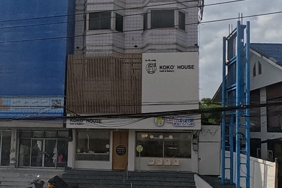 An old five-storey shophouse with a worn engraved sign on its parapet stands over ground-floor shops, including a clearly labelled KOKO' HOUSE Cafe & Bakery.