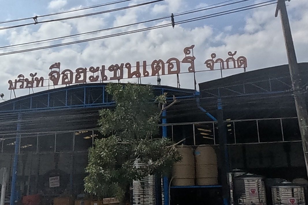 A large hardware and building-supplies warehouse named บริษัท จือฮะเซนเตอร์ จำกัด displays its name in big red letters above a yard stacked with water tanks and steel drums.