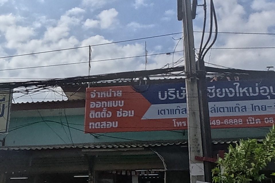 A yellow sign for ก๋วยเตี๋ยวเนื้อนายหมี noodle shop hangs beside a gas-appliance company's blue and orange sign advertising gas stoves, tanks and pipes with a phone number and opening hours; motorbikes are parked in the shopfront.