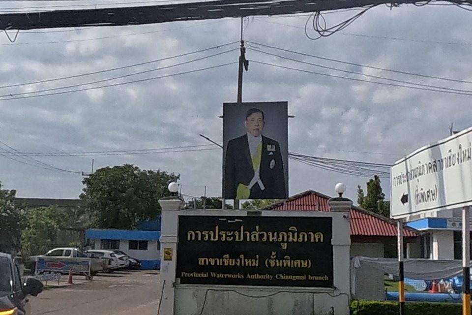 The gate of the Provincial Waterworks Authority, Chiang Mai branch (special class), shows its name in Thai and English on a sign board with a portrait of the King above, flags and hedges along the frontage, and roadworks with an excavator visible behind.