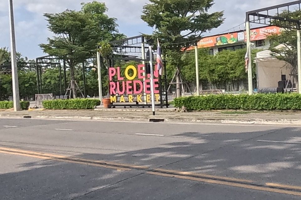 A colourful gateway sign reading PLOEN RUEDEE MARKET stands before a lively fenced market ground with tents, umbrellas and string lights.