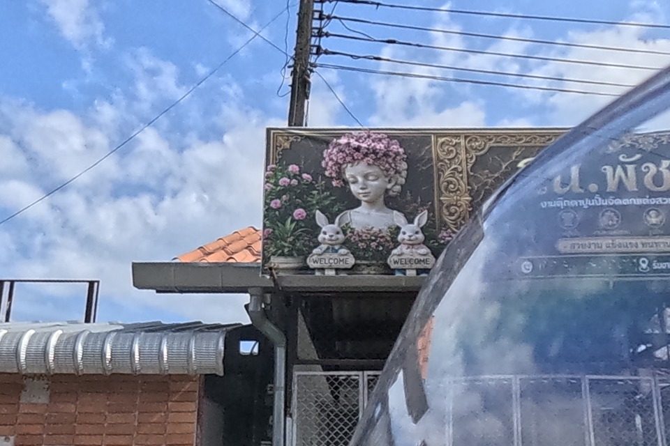 A garden-ornament shop sign shows a painted girl's bust crowned with flowers flanked by two cement rabbit statues holding 'WELCOME' plaques, gold scrollwork framing text reading 'ร้าน.พัชCha' with small service icons and a line 'concrete garden statue and decoration work, all kinds of jobs accepted'; a brick-faced building sits below.