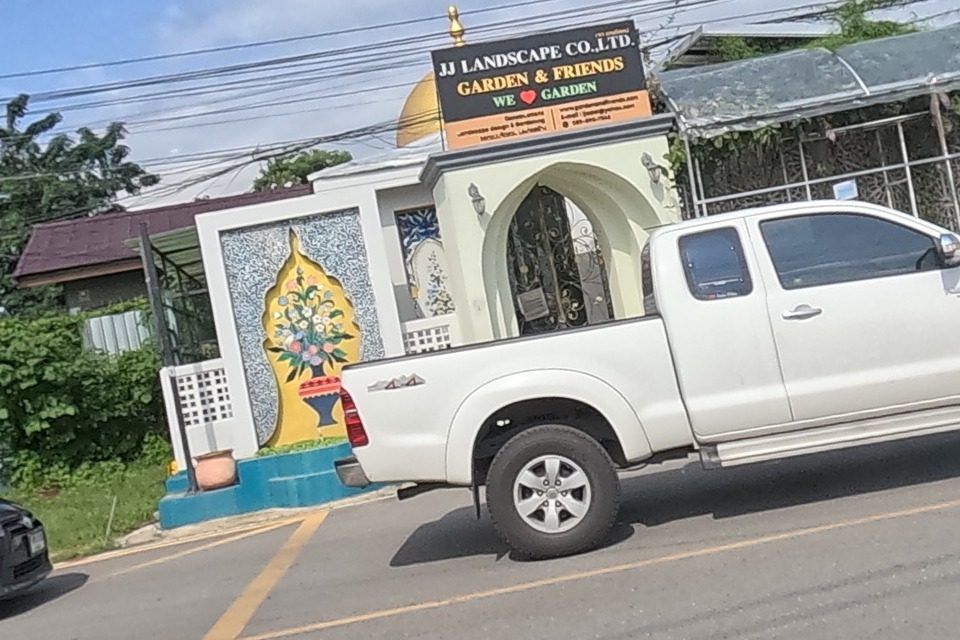 A decorative gateway reading 'JJ LANDSCAPE CO.,LTD — GARDEN & FRIENDS — WE (heart) GARDEN' with a gold dome and a mosaic mural of a flower vase; next to it, under a greenhouse roof, a garden-statue shop displays white cement figures with a sign 'Tel 053-131-788, 097-2393920' and a blank grey billboard frame stands behind; a white pickup and a black MPV pass on the road.