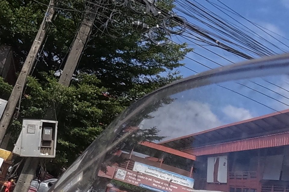 A parking area beside red-roofed wooden shophouses; a signboard on the upper floor names a church and a ground-floor board advertises motorbike, van and car trips to Pai.
