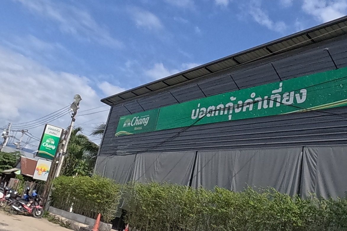 A black-clad warehouse-style building carries a green signboard reading 'บ่อตกกุ้งคำเที่ยง' next to a Chang mineral-water logo; motorbikes and several men are gathered outside.
