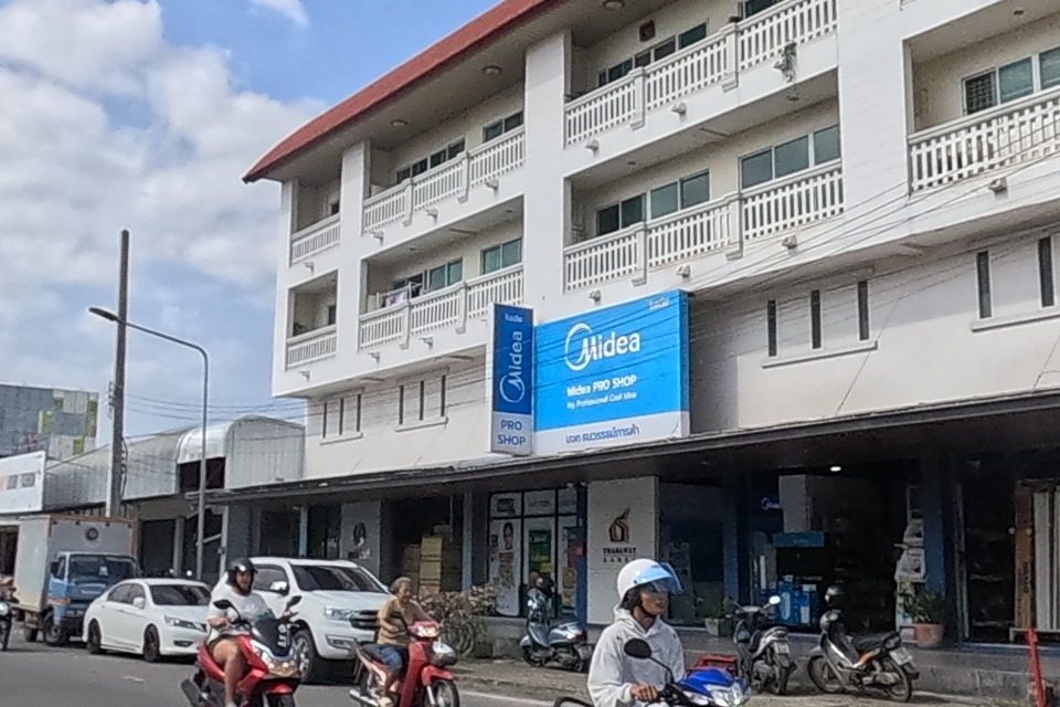 Across the road, a three-storey building with a Midea air-conditioner shop on the ground floor, and further right the หมิงยู่พลาสติก store again with a Falcon-brand sign.
