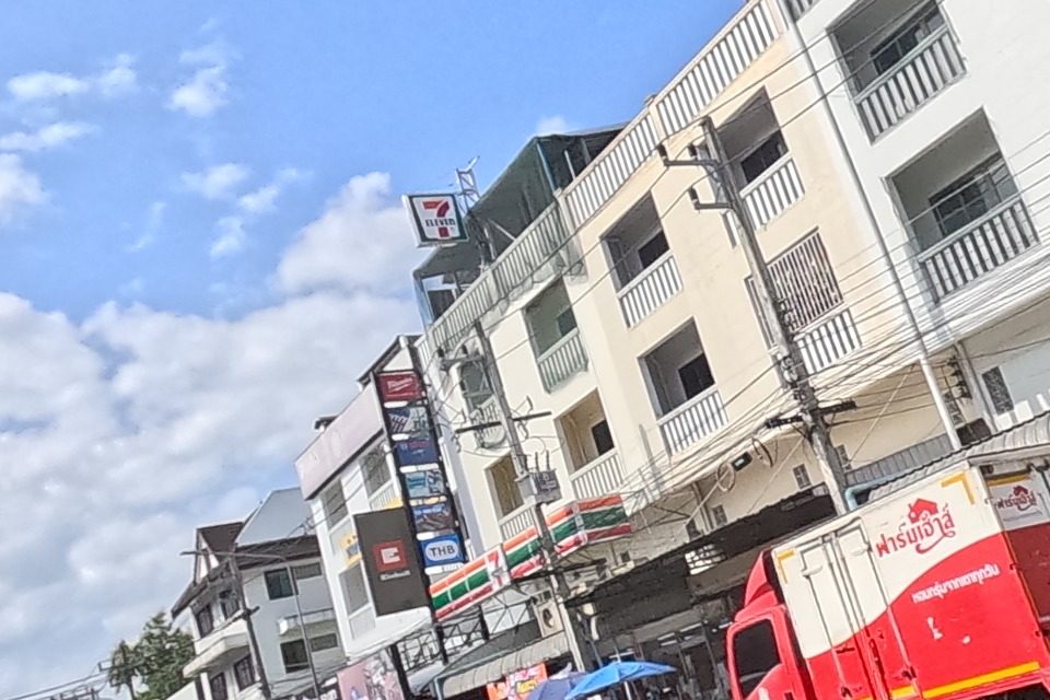 The road opens onto a wider junction where a 7-Eleven sign tops a shophouse block, with cars, mopeds and a street-food cart under a blue umbrella working the corner.
