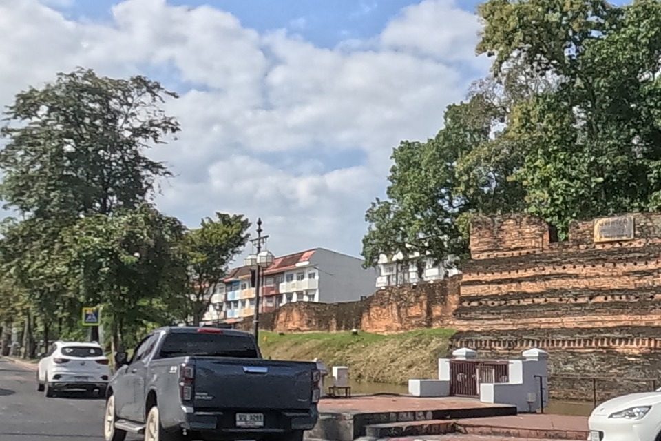 Heading toward the old city's northeast corner, the brick and white perimeter wall of the moat corner bastion (Chaeng Siphum) rises beyond a landscaped roundabout, with a Grab delivery rider and cars on the road.