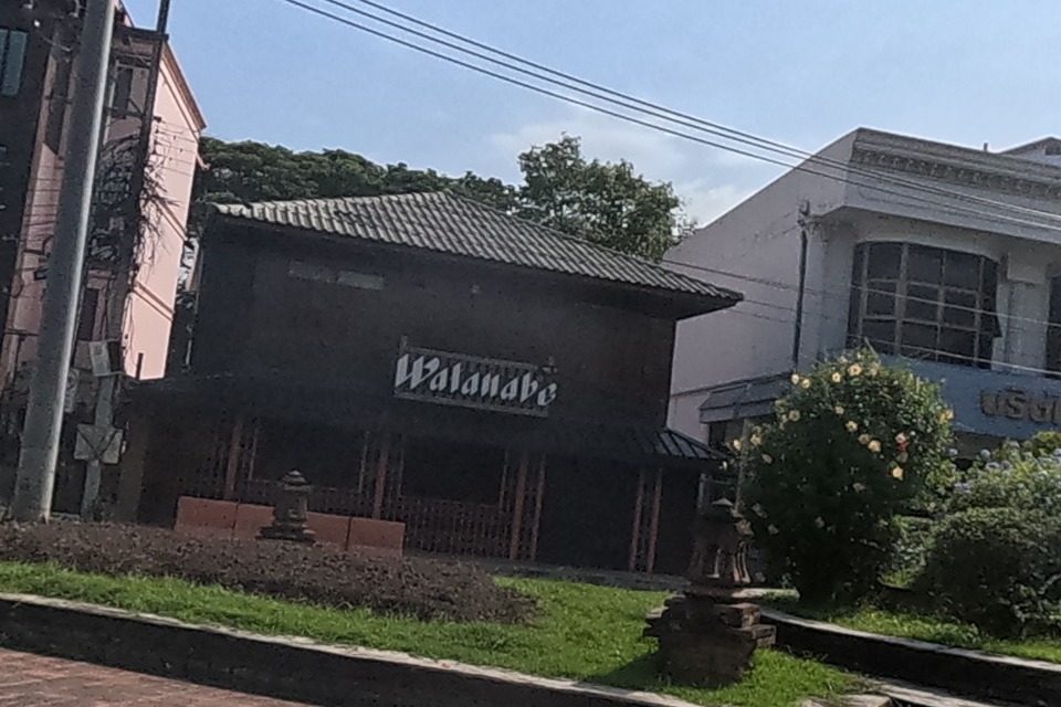 Watanabe, a Japanese restaurant in a traditional dark-teak building, stands beside a paved plaza with shrubs and a bare-brick corner bastion of the old city wall visible through the trees.