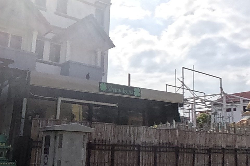 Shenanigans bar sign with a four-leaf-clover logo above a dark shopfront, with an ornate green-and-gold tiered Burmese/Shan-style temple roof visible further down the block.