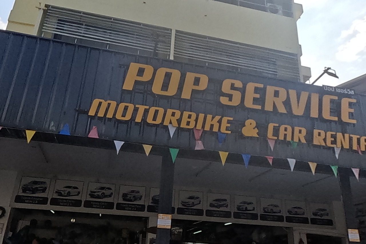 POP Service motorbike and car rental with a line of rental scooters out front, an ATM sign, and Ann's Rental bike-and-car-for-rent across the side street with a yellow scooter parked.
