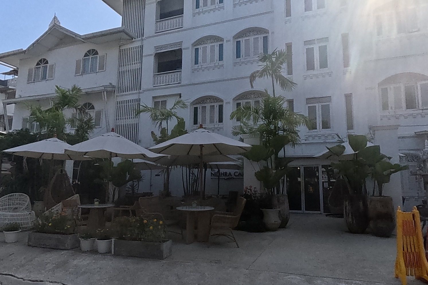 Nidhra Lanna Hotel's white colonial-style facade with outdoor cafe seating under umbrellas, a tour-information board, and a Thai spirit house in shadow by the entrance.